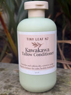 A bottle of Tiny Leaf NZ Kawakawa Tallow Conditioner lying diagonally on a textured stone surface with blurred natural foliage in the background.