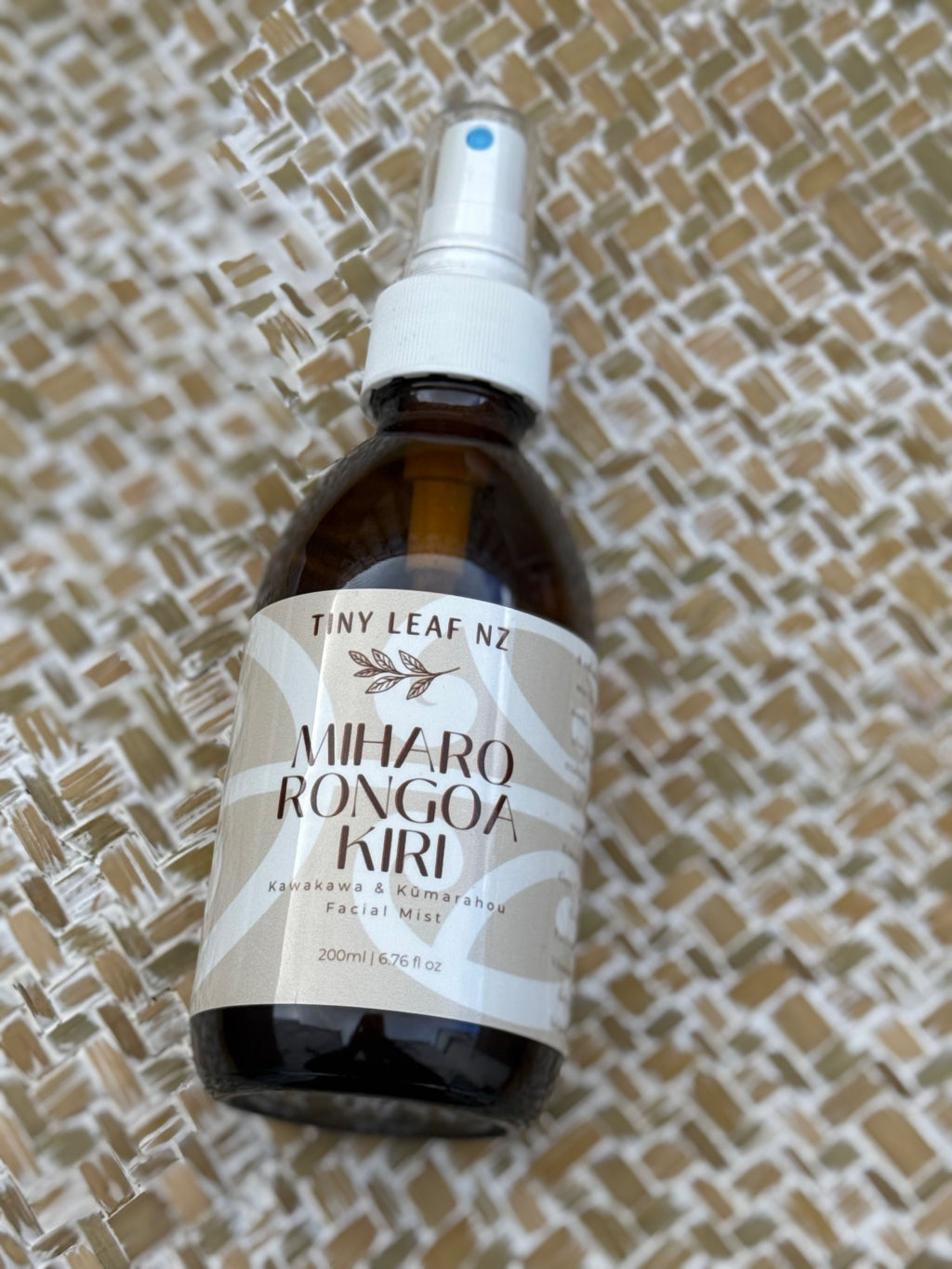 A 200ml amber glass spray bottle of Miharo Rongoa Kiri Facial Mist by Tiny Leaf Nz, featuring a white spray nozzle and a beige and white label, resting on a woven straw background.