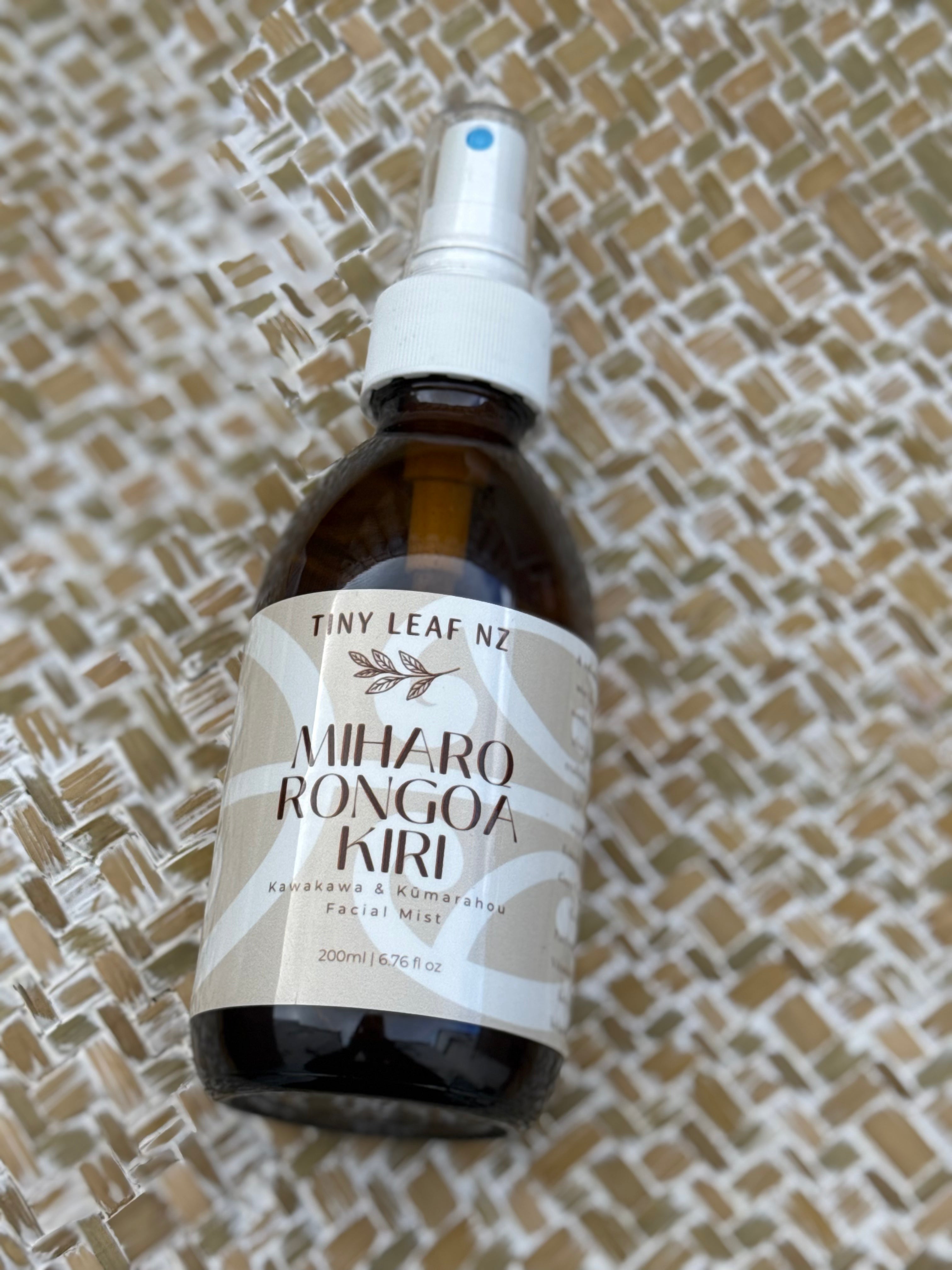 A 200ml amber glass spray bottle of Miharo Rongoa Kiri Facial Mist by Tiny Leaf Nz, featuring a white spray nozzle and a beige and white label, resting on a woven straw background.