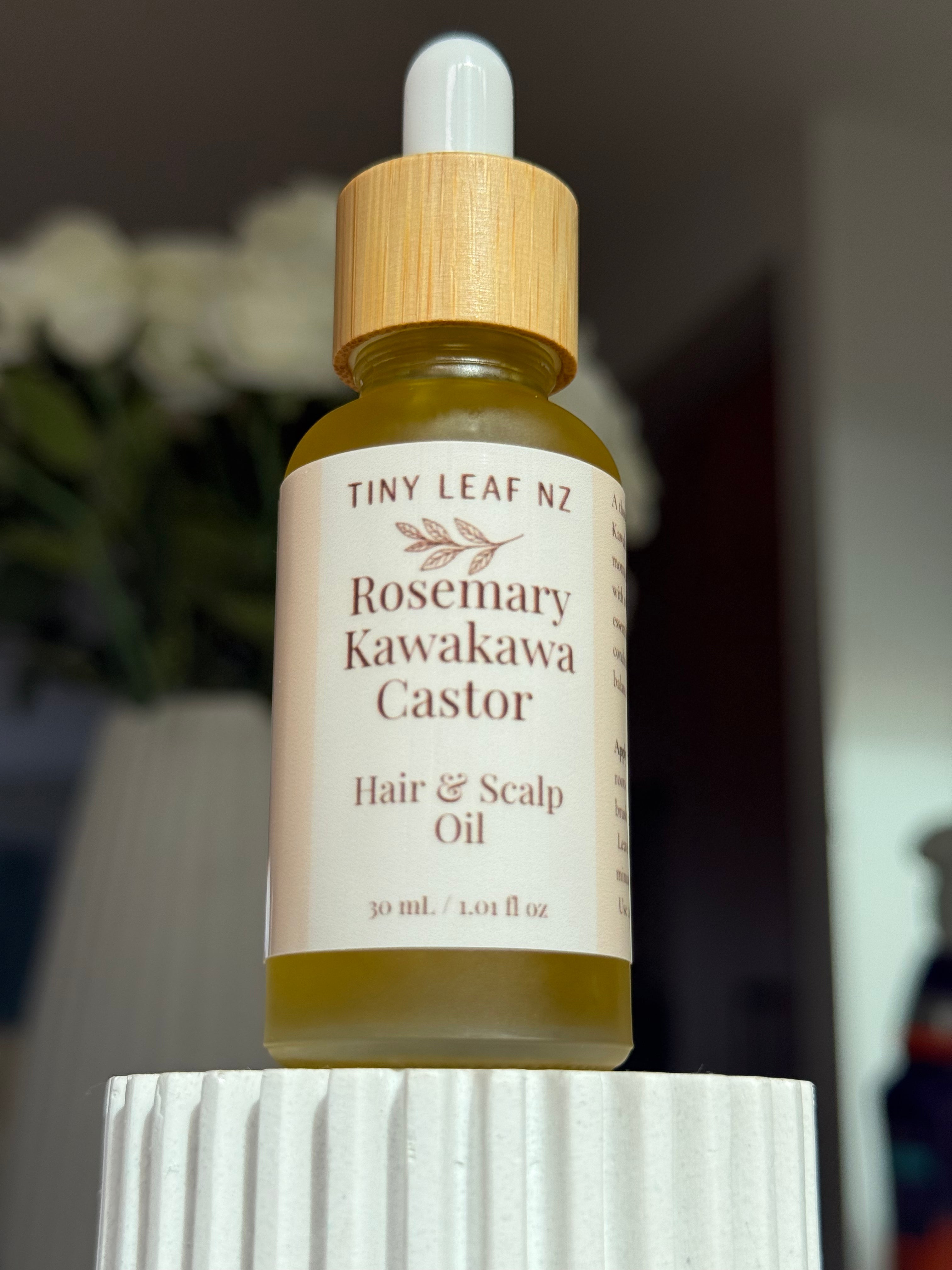 A bottle of Tiny Leaf NZ Rosemary Kawakawa Castor Hair & Scalp Oil, featuring a light wood dropper cap and a cream-colored label, displayed on a white ribbed pedestal with a blurred indoor background.