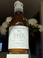 A bottle of Tiny Leaf Nz Kawakawa Oat Daily Lotion, 300 ml, featuring an amber brown bottle and a white label, displayed on a white pedestal with blurred white flowers in the background.