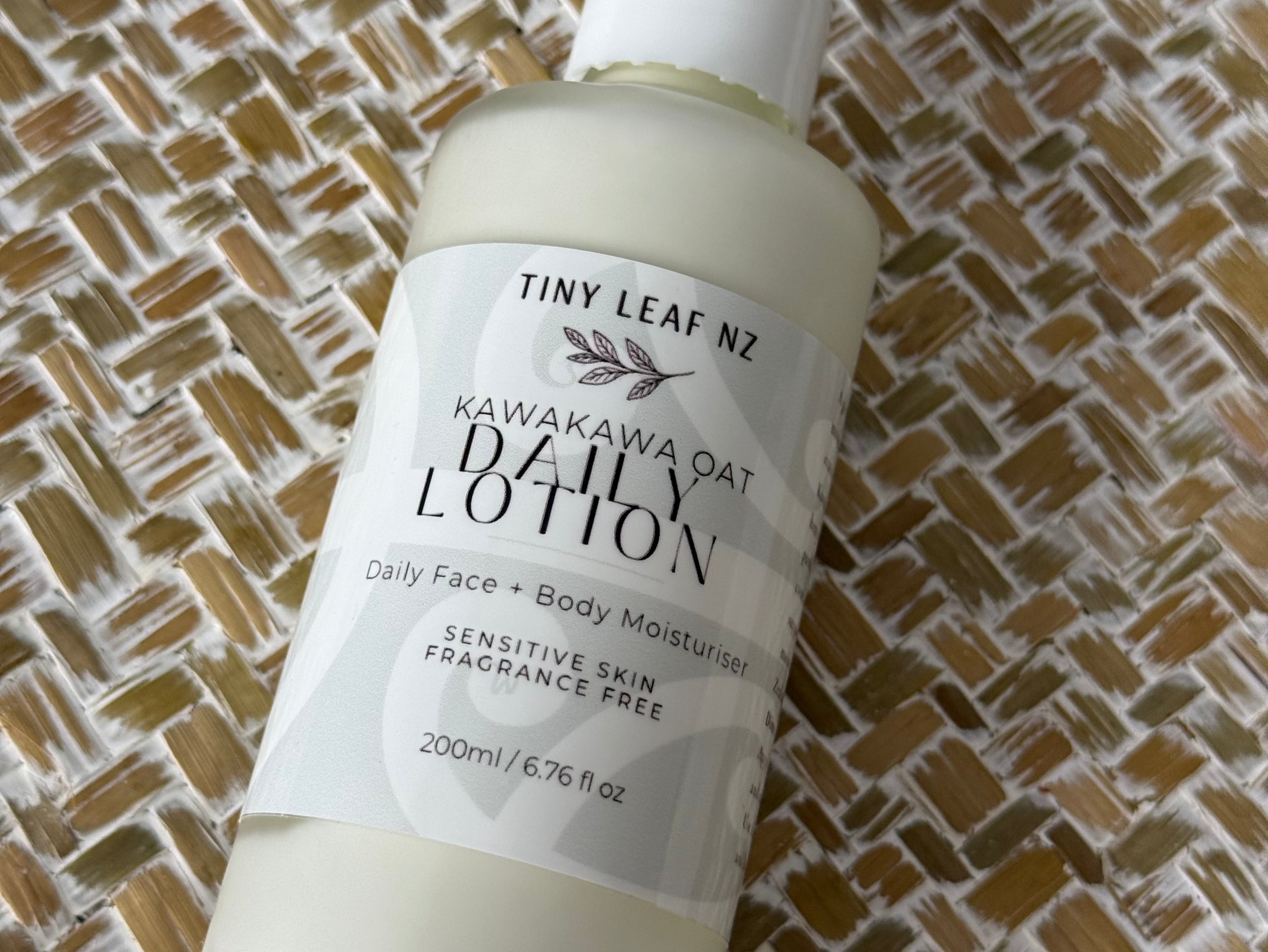 A bottle of Tiny Leak NZ Hawaiian Oat Body Lotion with a white pump dispenser, lying diagonally on a woven brown and beige textured mat.