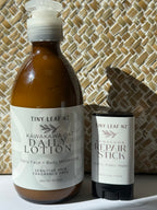 A bottle of Tiny Leaf NZ Kawakawa Oat Daily Lotion next to a matching stick of Tiny Leaf NZ Kawakawa Repair Stick, set against a woven straw background.