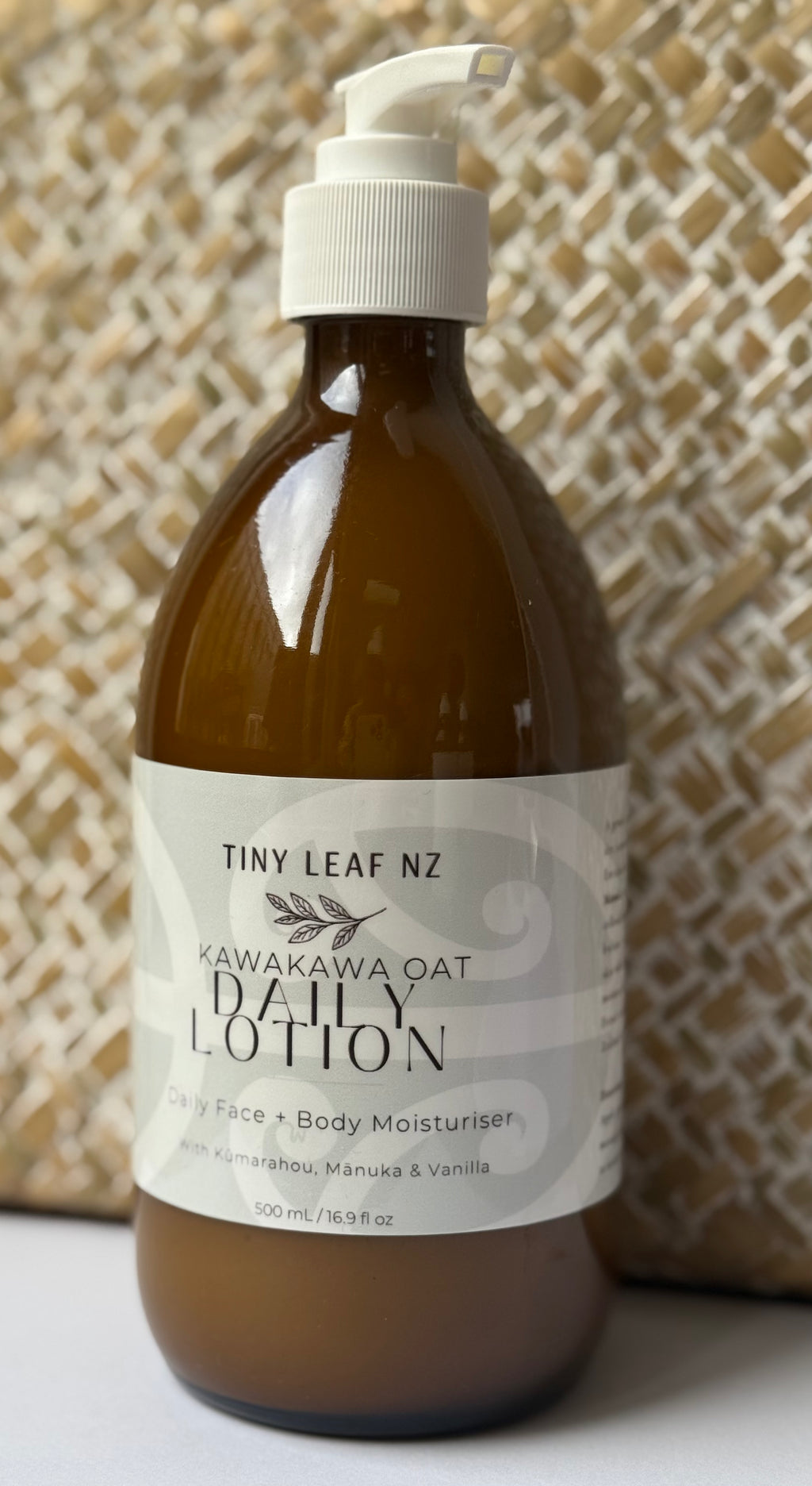 A 500ml pump bottle of Tiny Leaf NZ Kawakawa Oat Daily Lotion, made of amber brown glass with a white pump and a white label, set against a woven beige background.