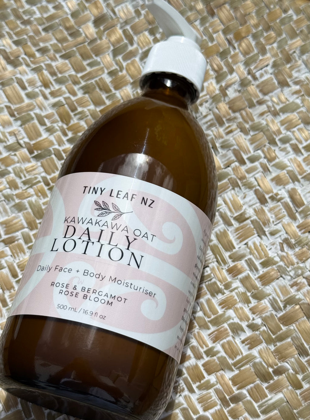 A bottle of Tiny Leaf Nz Kawakawa Oat Daily Lotion with a white pump dispenser and a pink and white label, resting on a woven straw mat background.