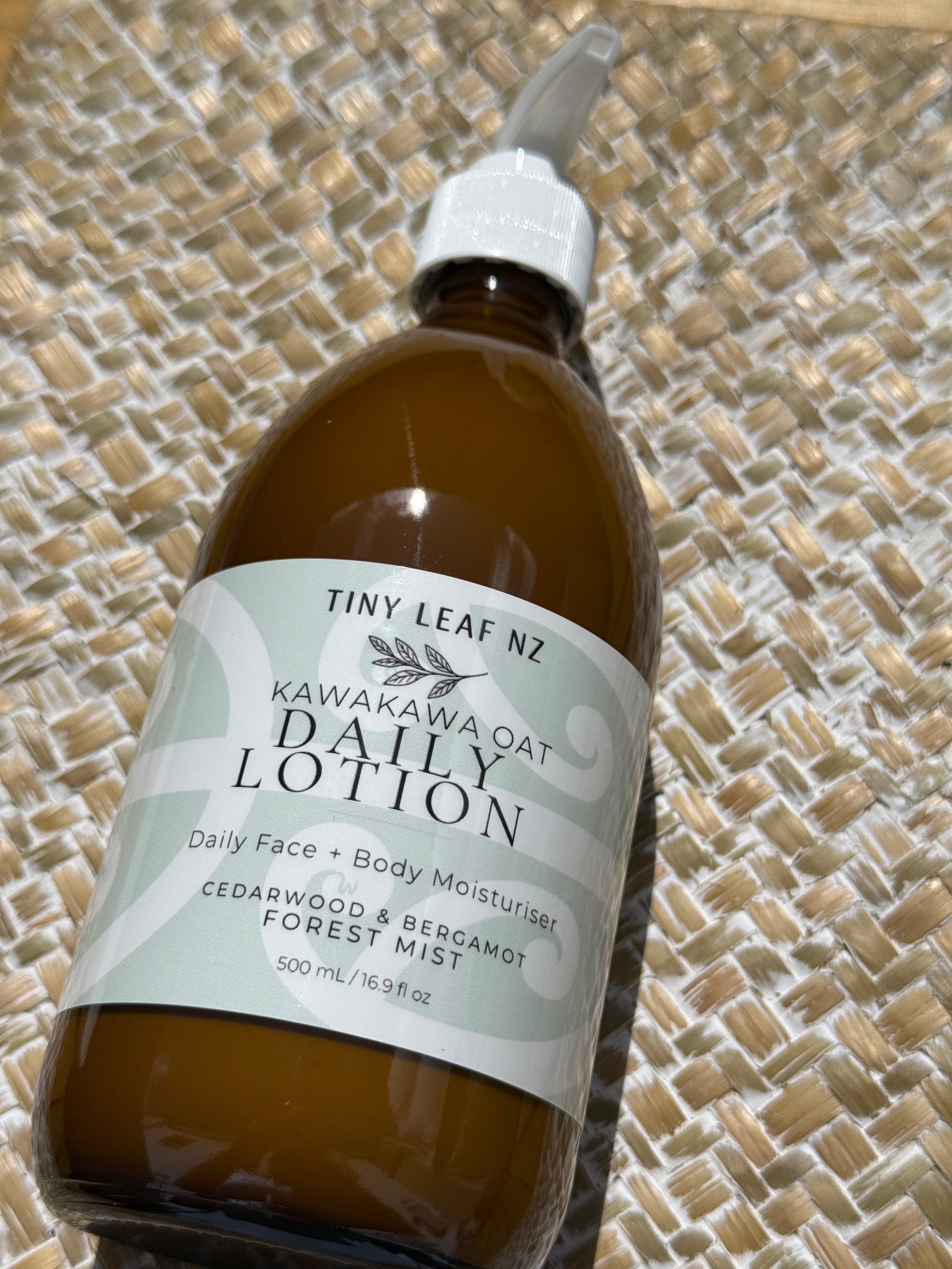 A 500ml amber glass bottle of Tiny Leaf Nz Kawakawa Oat Daily Lotion with a white spray cap, resting on a woven straw mat.