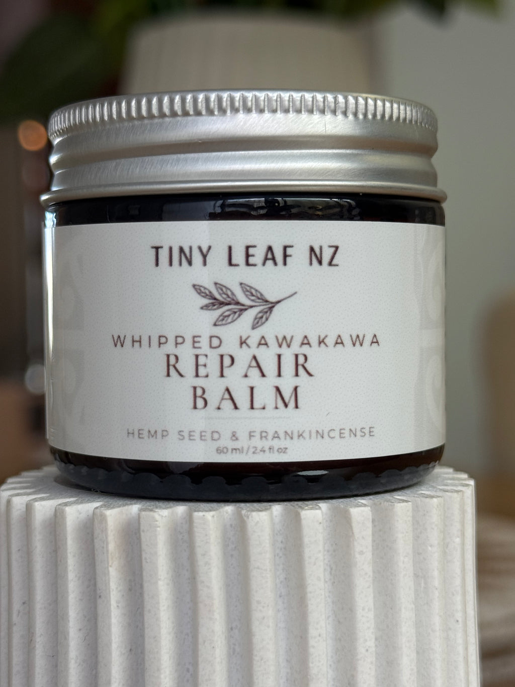 A jar of Tiny Leaf NZ Whipped Kawakawa Repair Balm with a white label and silver lid, displayed on a white ribbed pedestal.