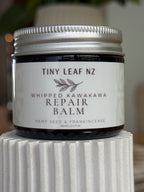A jar of Tiny Leaf NZ Whipped Kawakawa Repair Balm with a white label and silver lid, displayed on a white ribbed pedestal.