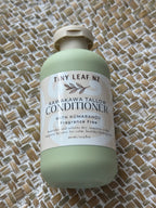 A bottle of Tiny Leaf Nz Kawakawa Tallow Conditioner with a pale sage green body and cream-colored cap, resting on a woven straw mat background.