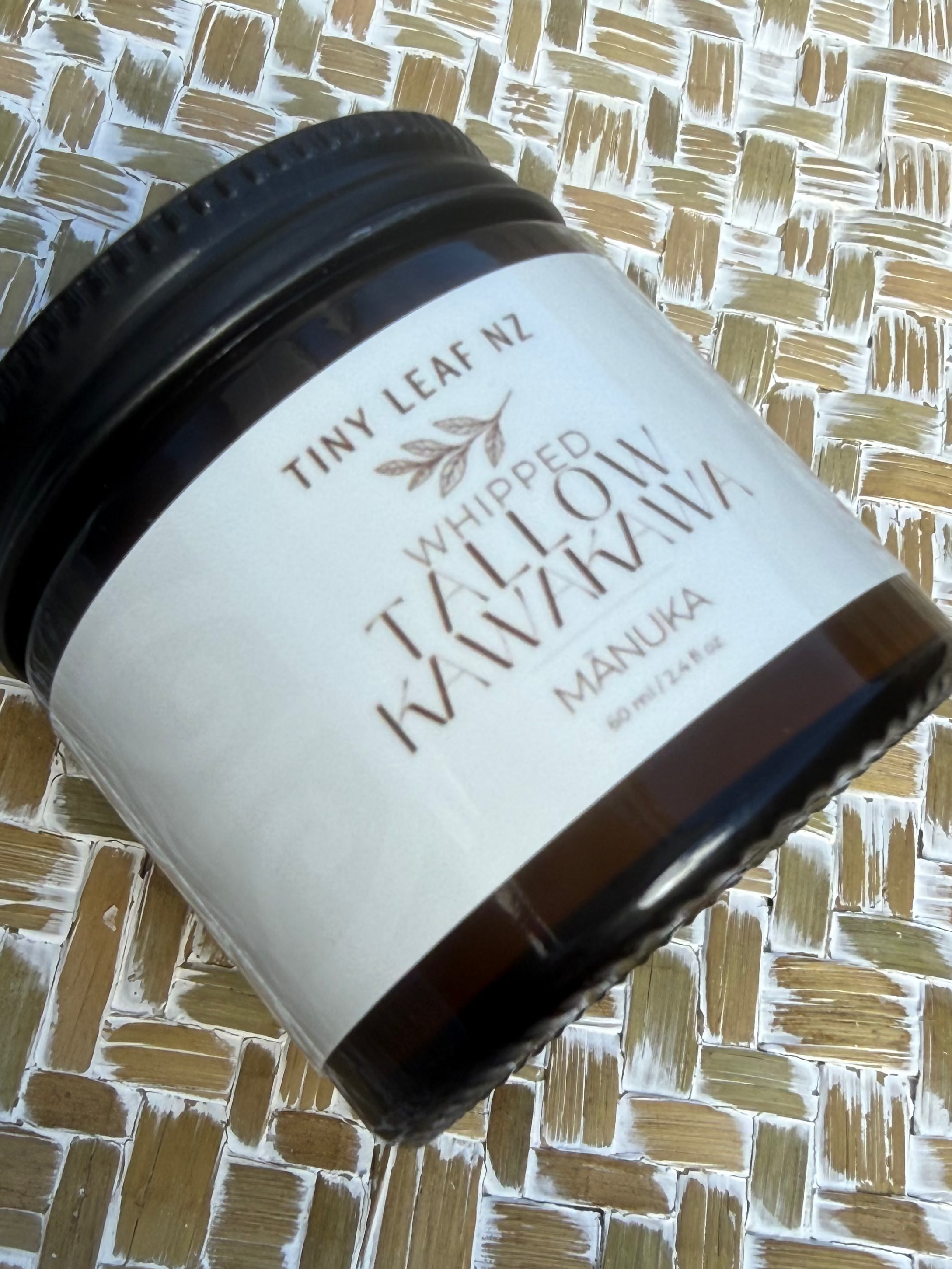 A dark amber glass jar of Tiny Leaf N2 Whipped Tallow Mavimava Manuka, featuring a white label with brown text, resting on a textured woven mat.
