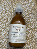 A bottle of Tiny Leaf Nz Kawakawa Tallow Conditioner with Kumarahou, placed on a woven straw mat background.