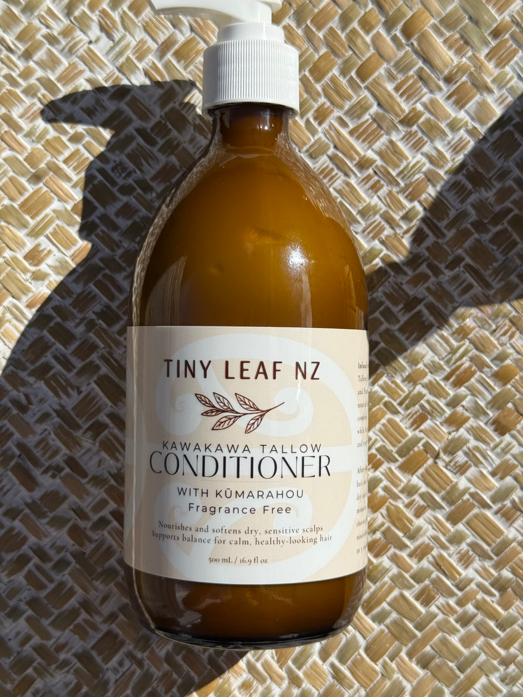 A bottle of Tiny Leaf Nz Kawakawa Tallow Conditioner with Kumarahou fragrance free, resting on a woven straw mat under direct sunlight.
