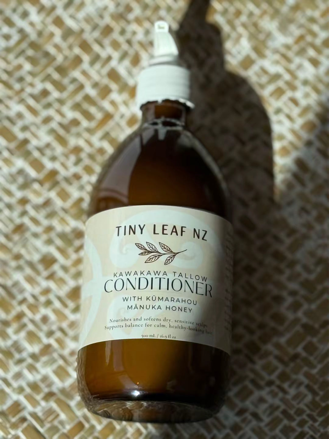 A bottle of Tiny Leaf NZ Kawakawa Tallow Conditioner with Kumarahou Manuka Honey, featuring an amber glass bottle and a cream-colored label, set against a woven straw background.