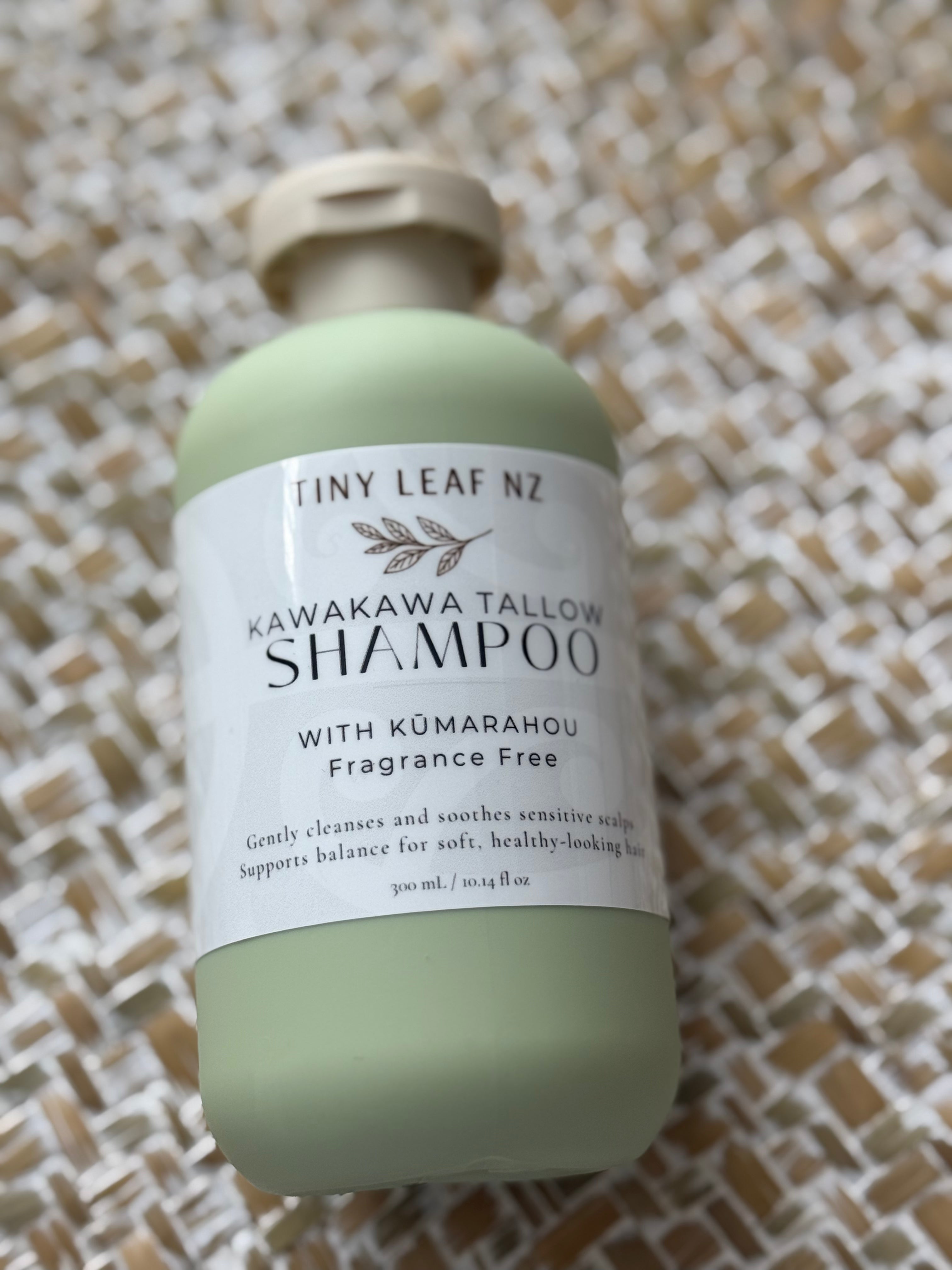 A bottle of Tiny Leaf NZ Kawakawa Tallow Shampoo with a pale sage green bottle and a white label, resting on a woven beige and white mat.