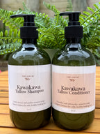 Two bottles of Kawakawa Tallow Shampoo and Conditioner on a wooden surface with green leaves in the background.