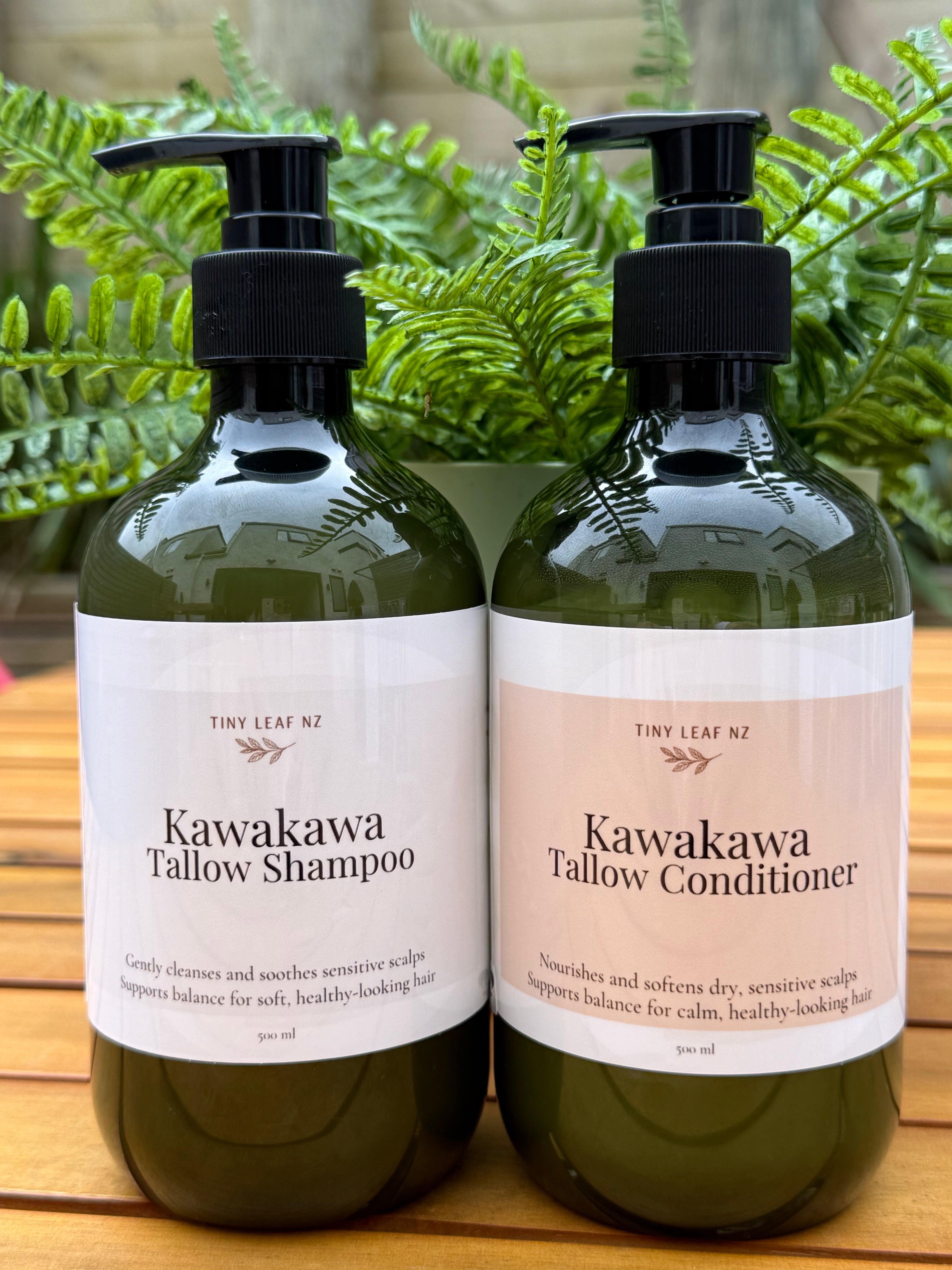 Two bottles of Kawakawa Tallow Shampoo and Conditioner on a wooden surface with green leaves in the background.