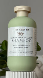 A bottle of Tiny Leaf NZ Kawakawa Tallow Shampoo with Kumerahu, in a pale sage green bottle with a cream-colored pump and a white label, set against a neutral gray background with soft green leaves in the background.