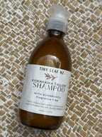 A bottle of Tiny Leaf Nz Kawakawa Tallow Shampoo with Kumarahou Fragrance Free, resting on a woven straw mat background.