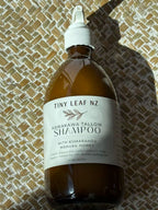 A bottle of Tiny Leaf Nz Kawakawa Tallow Shampoo resting on a woven straw mat under direct sunlight. The bottle is amber brown with a white cap and a white label.