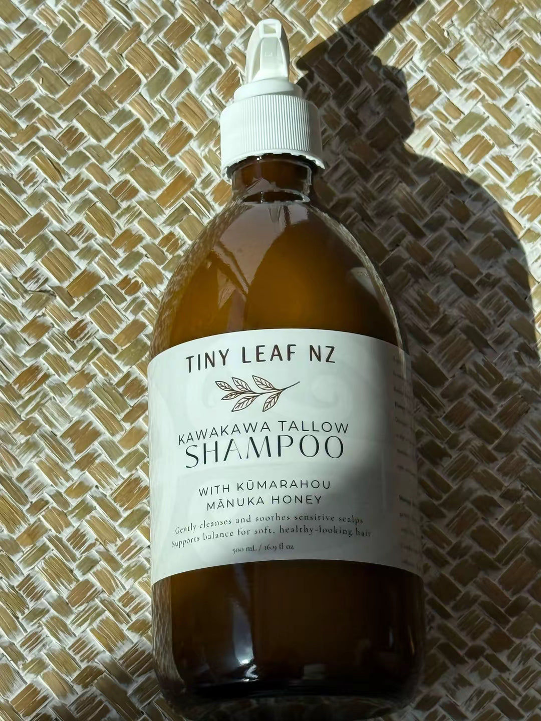 A bottle of Tiny Leaf Nz Kawakawa Tallow Shampoo resting on a woven straw mat under direct sunlight. The bottle is amber brown with a white cap and a white label.