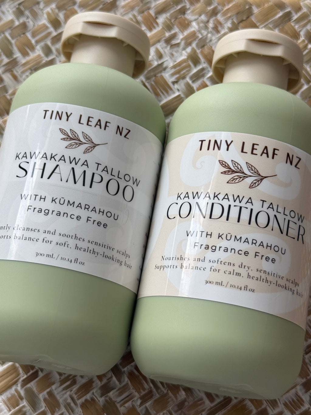 Two pale sage green bottles of Tiny Leaf Nz hair care products, labeled Kawakawa Tallow Shampoo and Kawakawa Tallow Conditioner, placed on a woven mat background.