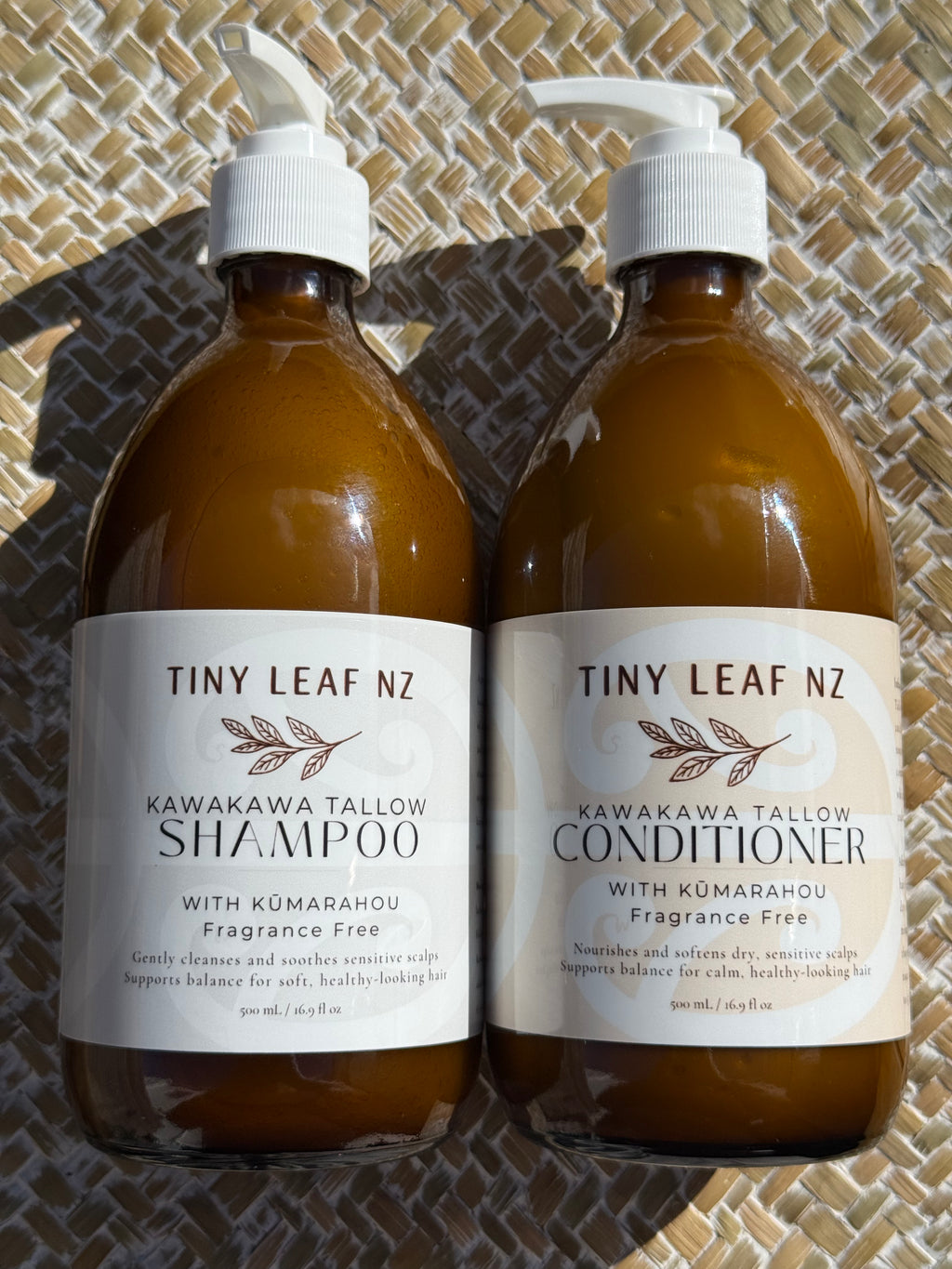 Two amber glass pump bottles of Tiny Leaf Nz haircare products, labeled Kawakawa Tallow Shampoo and Kawakawa Tallow Conditioner, placed side by side on a woven mat background.
