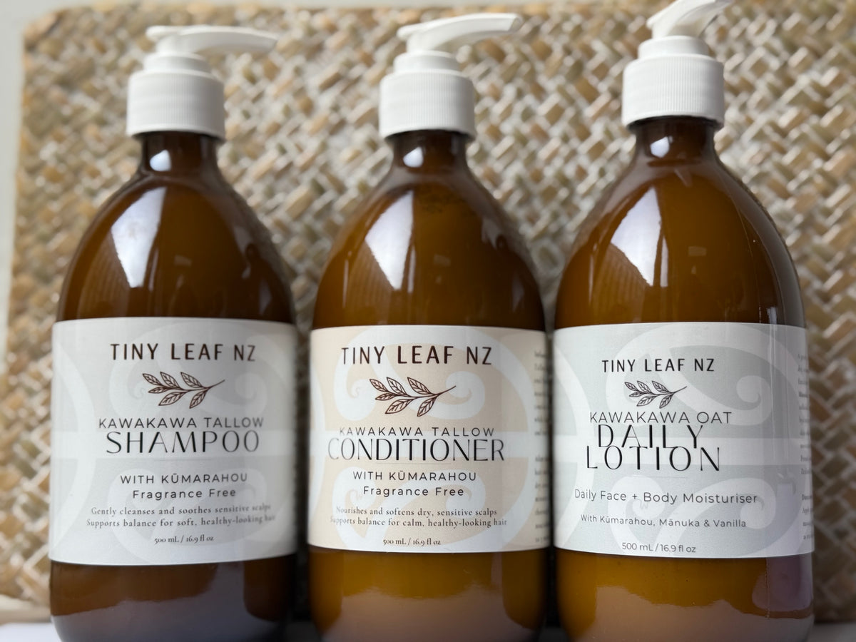 Three amber plastic pump bottles of Tiny Leaf NZ haircare products lined up against a woven beige background: Kawakawa Tallow Shampoo, Kawakawa Tallow Conditioner, and Kawakawa Oat Daily Lotion.