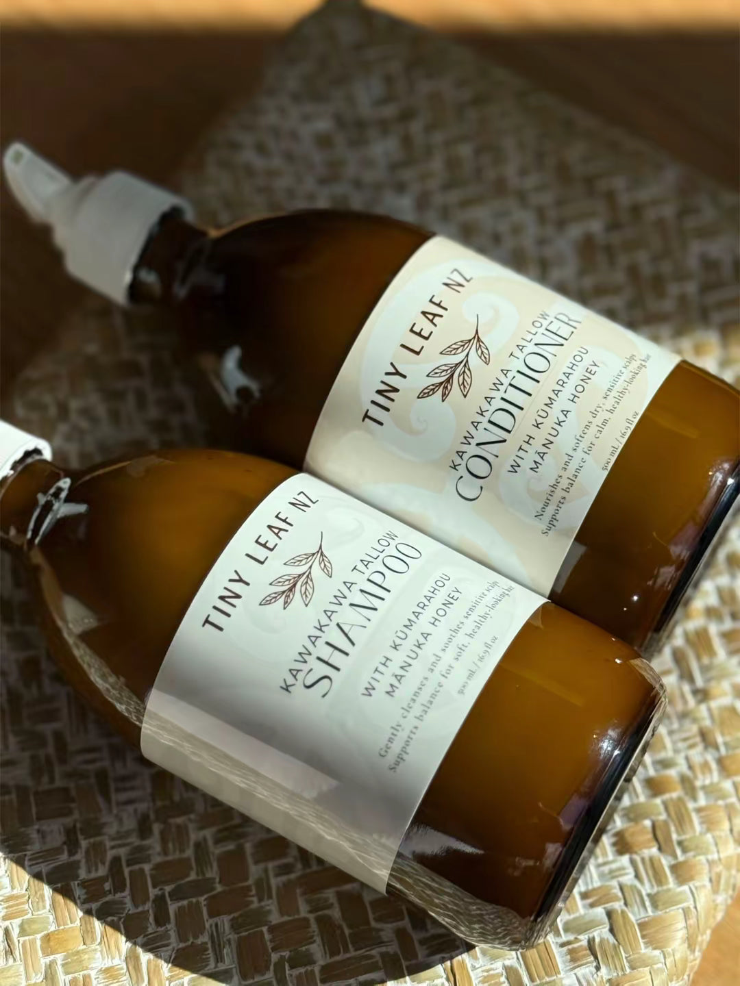 Two amber glass bottles of Tiny Leaf shampoo and conditioners with white labels, resting on a woven straw mat.