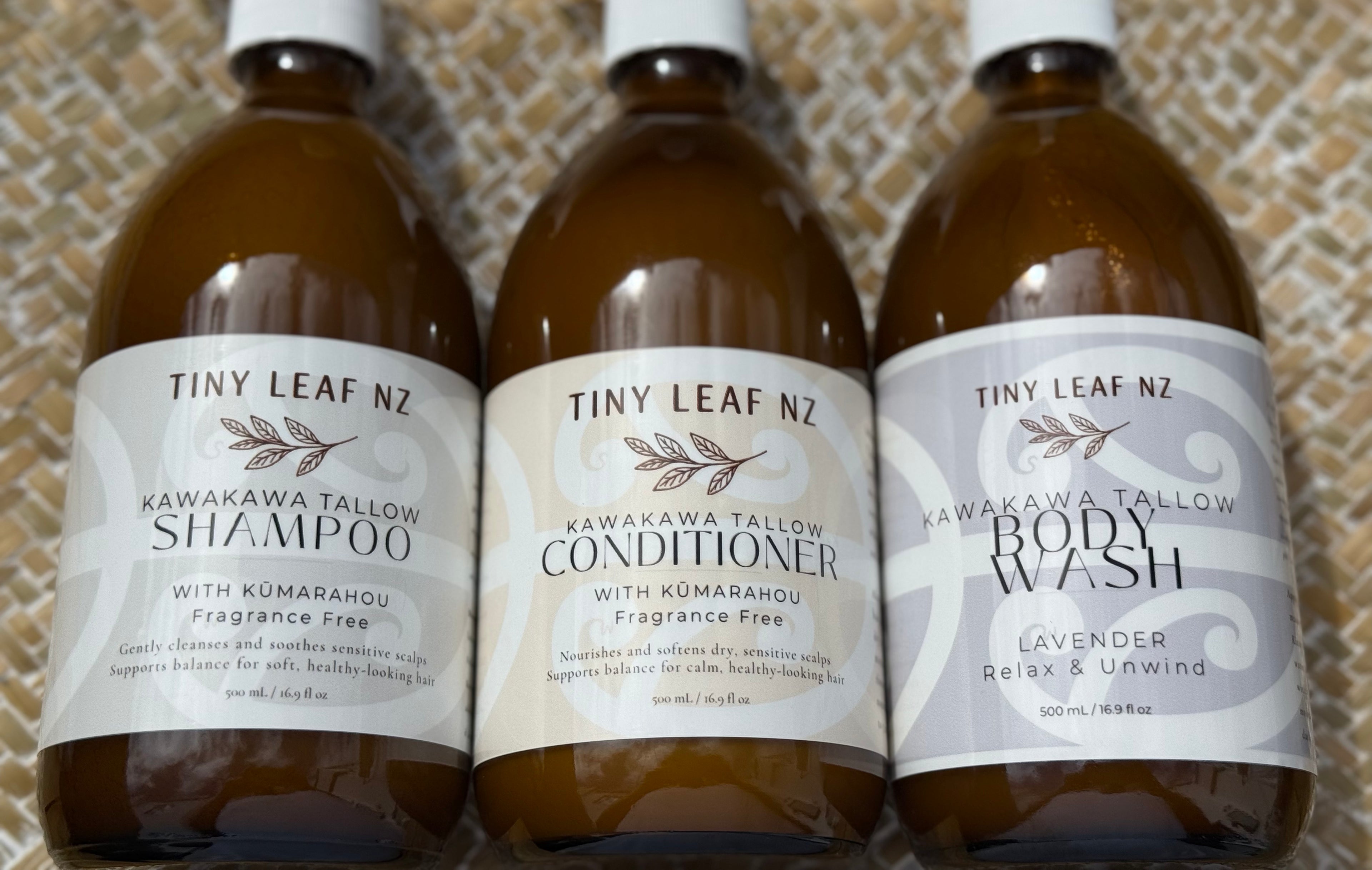 Three amber glass bottles of Tiny Leaf Nz Kawakawa Tallow Care products lined up on a textured beige woven mat: Shampoo, Conditioner, and Body Wash.