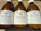 Three amber plastic bottles of Tiny Leaf NZ hair and skin care products lined up on a woven mat background: Kawakawa Tallow Shampoo, Kawakawa Tallow Conditioner, and Kawakawa Daily Lotion.