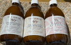 Three amber plastic bottles of Tiny Leaf Nz haircare products lined up on a patterned beige surface: Kawakawa Tallow Shampoo, Kawakawa Tallow Conditioner, and Kawakawa Cat Daily Lotion.