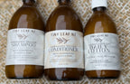 Three amber glass bottles of Tiny Leaf NZ hair care products lined up on a textured beige woven mat: Shampoo, Conditioner, and Daily Lotion.