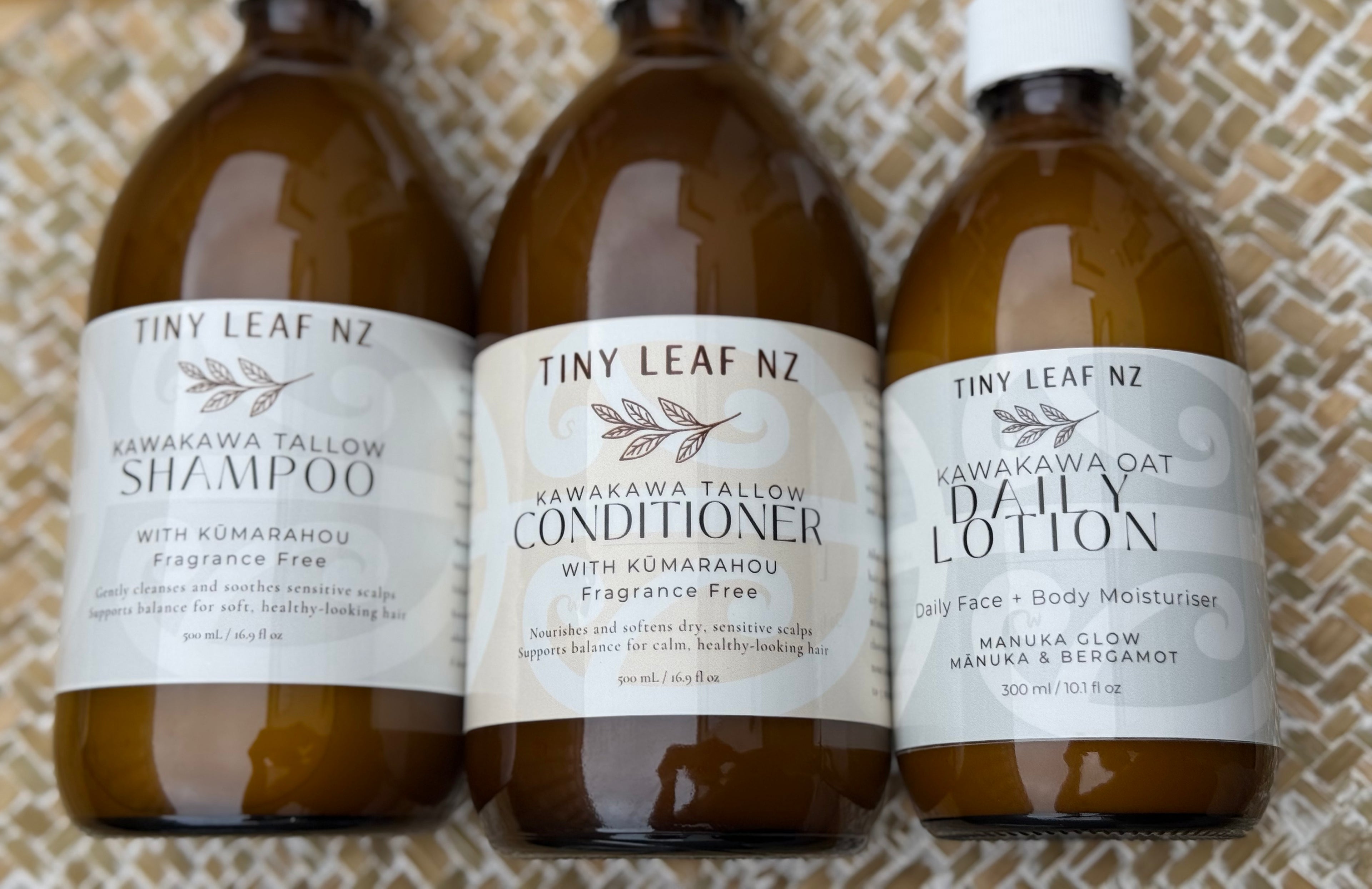 Three amber glass bottles of Tiny Leaf NZ hair care products lined up on a textured beige woven mat: Shampoo, Conditioner, and Daily Lotion.