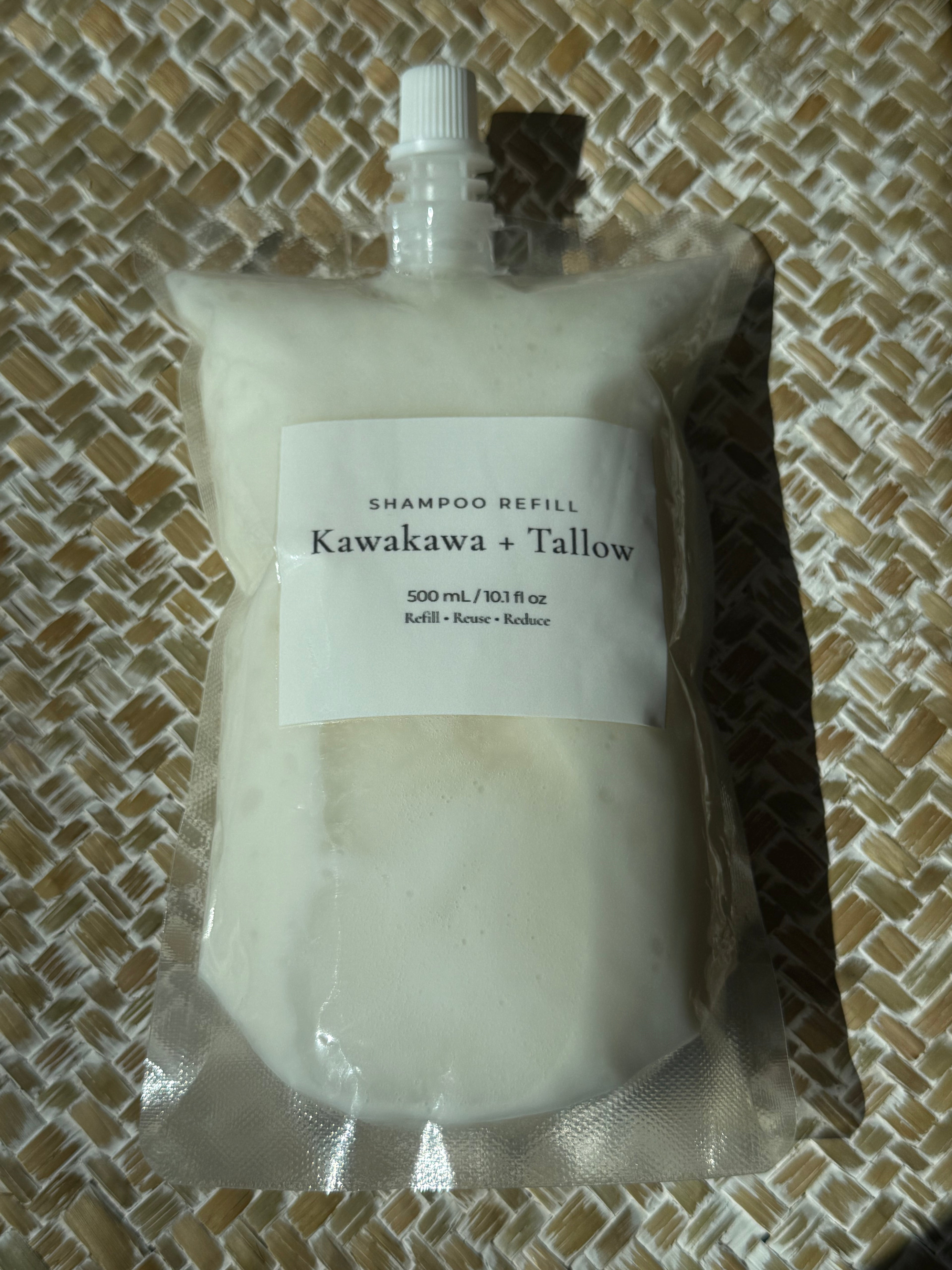 A 500 mL shampoo refill pouch labeled 'Kawakawa + Tallow' filled with off-white cream, sealed in a clear plastic bag with a white spout cap, placed on a woven beige mat.