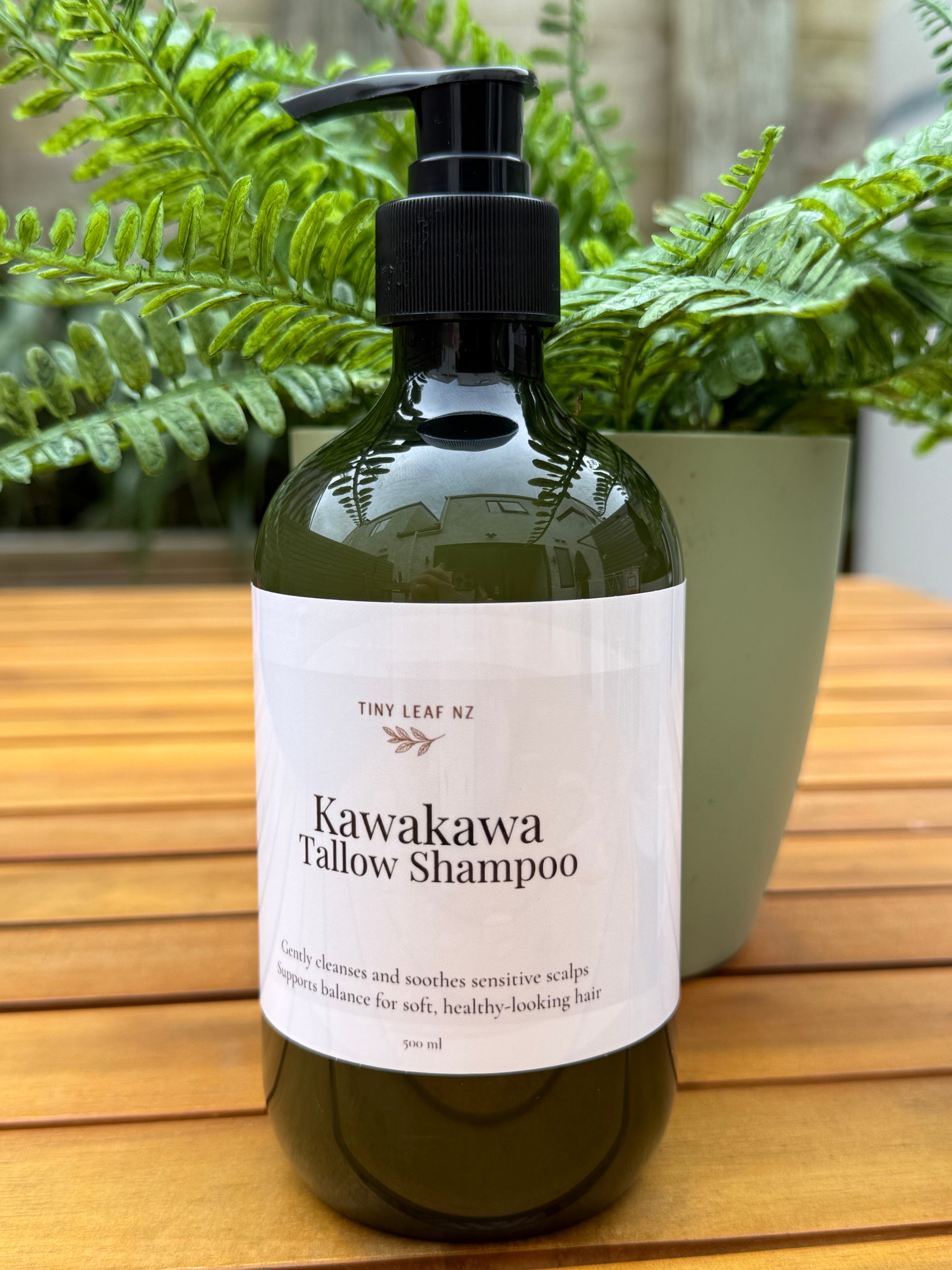 Bottle of Kawakawa Tallow Shampoo on a wooden surface with green plants in the background