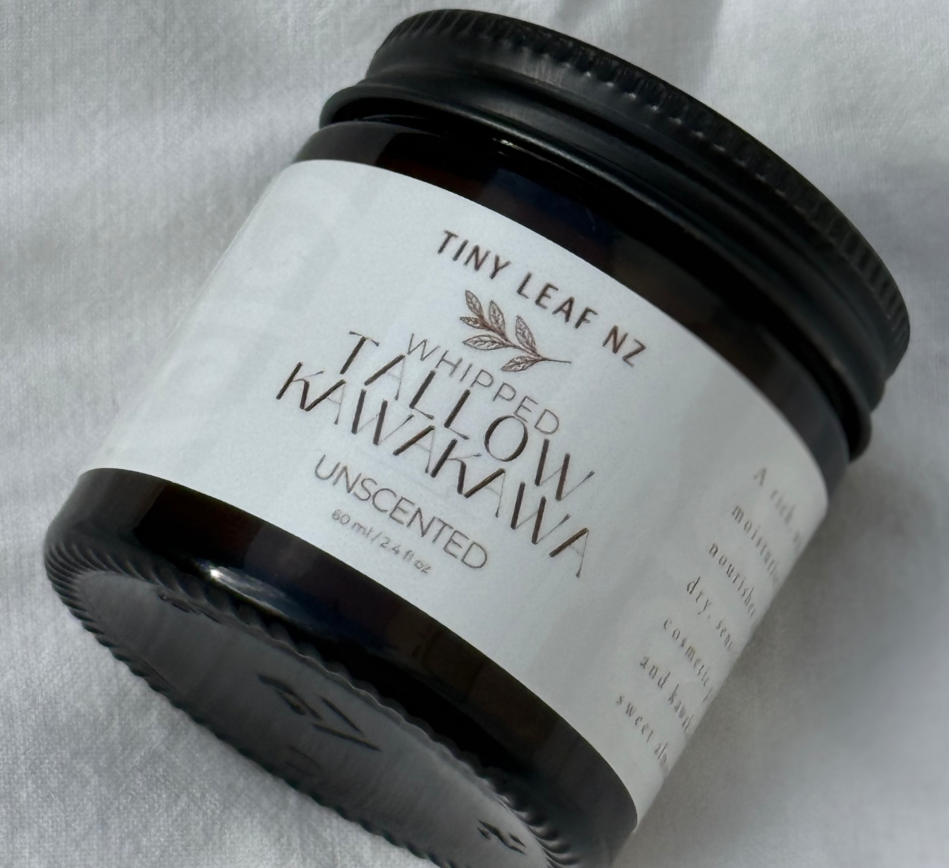 A small black jar of Tiny Leaf NZ Whipped Waxowhaukawa Unscented wax, shown on a textured white fabric background.