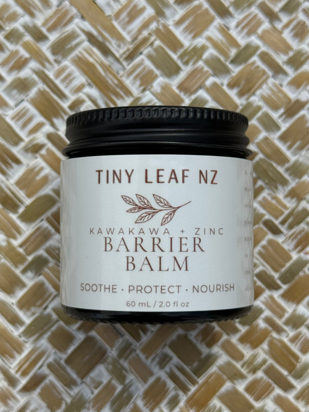 A jar of Tiny Leaf NZ Kawakawa + Zinc Barrier Balm with a black lid and white label, placed on a woven straw mat background.