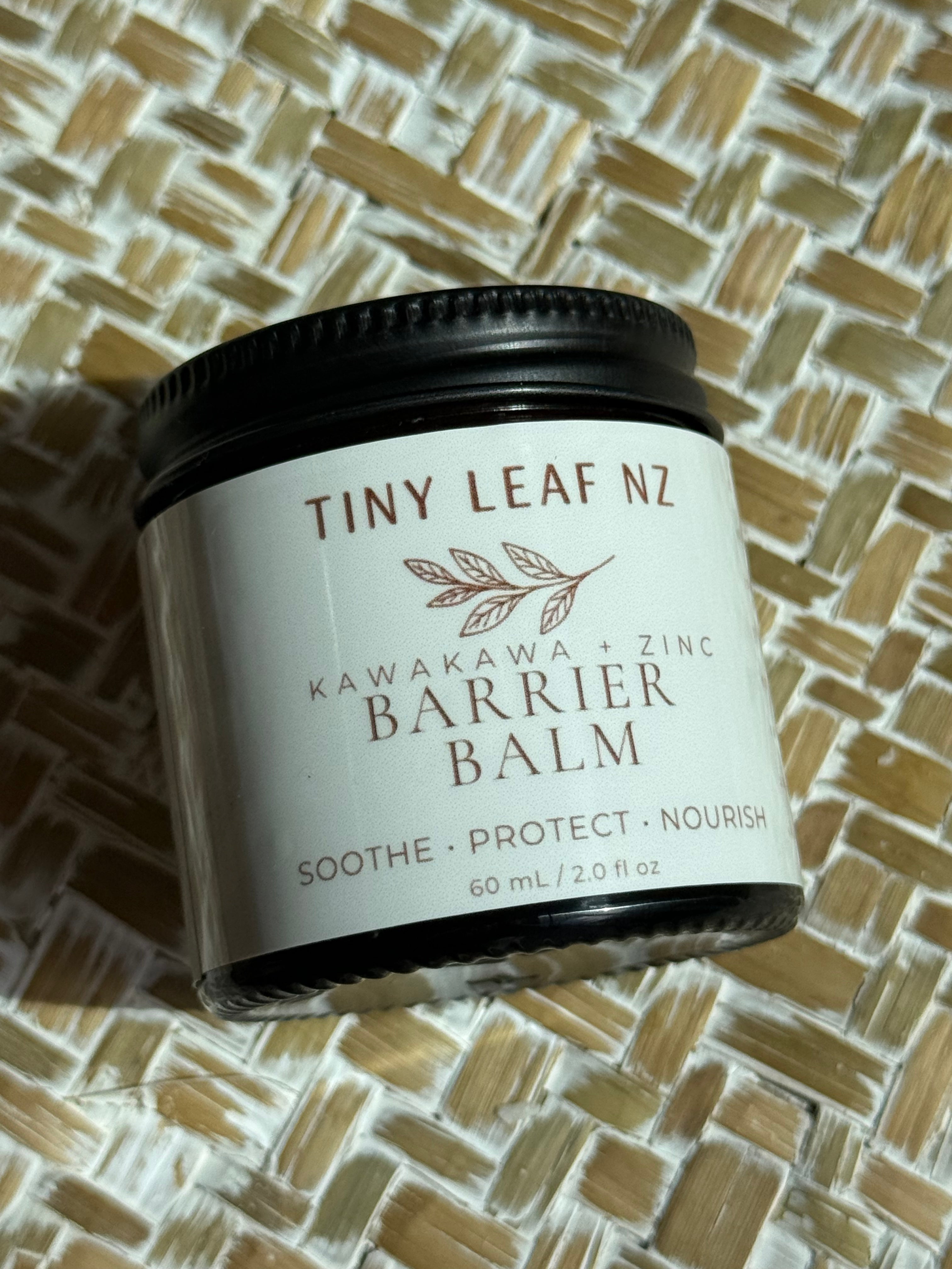 A small jar of Tiny Leaf Nz Kawakawa + Zinc Barrier Balm resting on a woven straw mat. The jar has a white label with brown text and a dark brown lid.