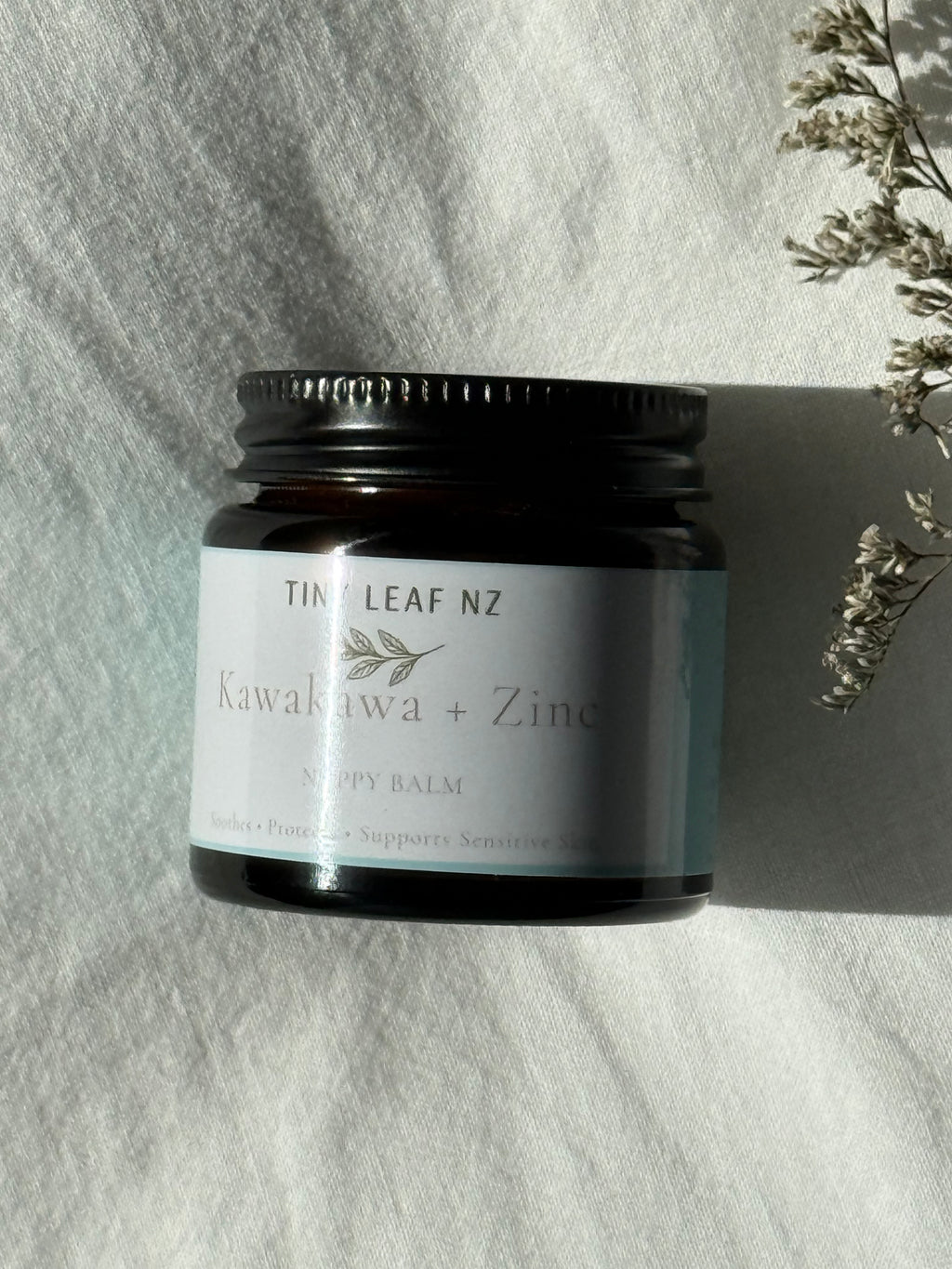 Tin of Tiny leaf NZ balm on a textured white surface with dried flowers.