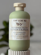 A bottle of Tiny Leaf Nz Kawakawa Tallow Conditioner with Kumerahou, in a sage green bottle with a cream-colored cap and label, displayed on a white ribbed pedestal against a soft gray background.