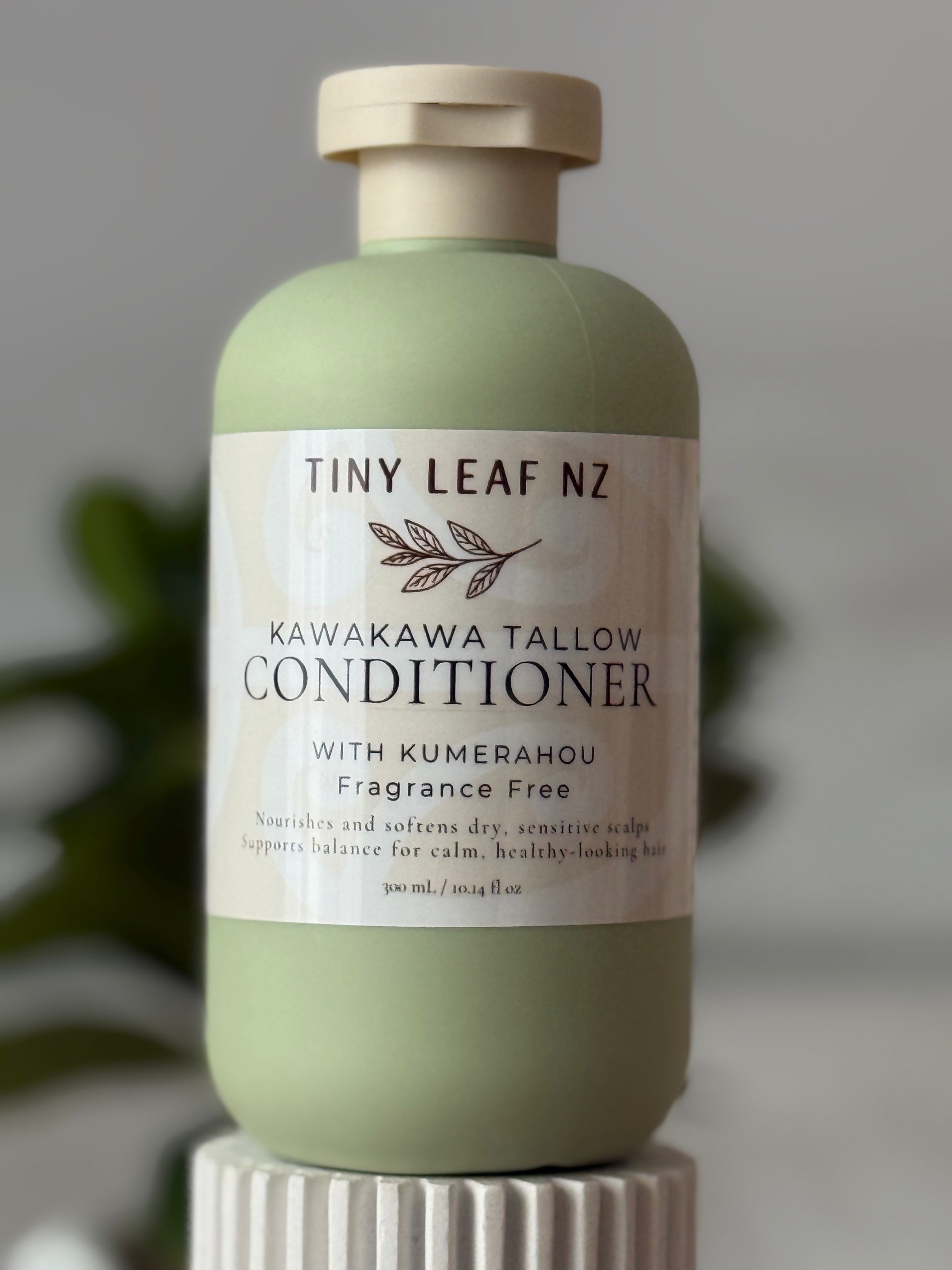 A bottle of Tiny Leaf Nz Kawakawa Tallow Conditioner with Kumerahou, in a sage green bottle with a cream-colored cap and label, displayed on a white ribbed pedestal against a soft gray background.