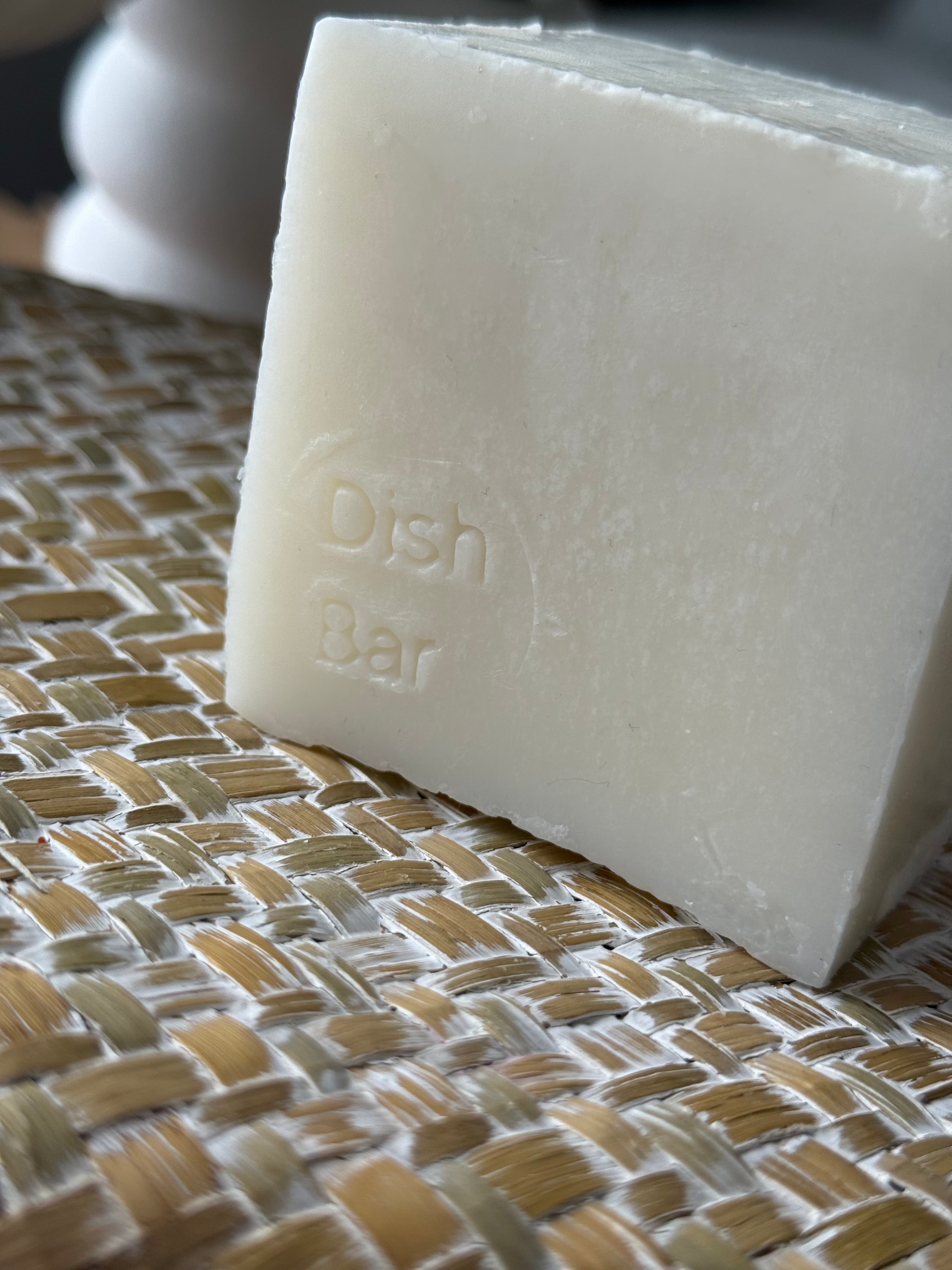 A close-up of a rectangular bar of off-white dish soap embossed with the words 'Dish Bar', resting on a woven beige and white placemat.