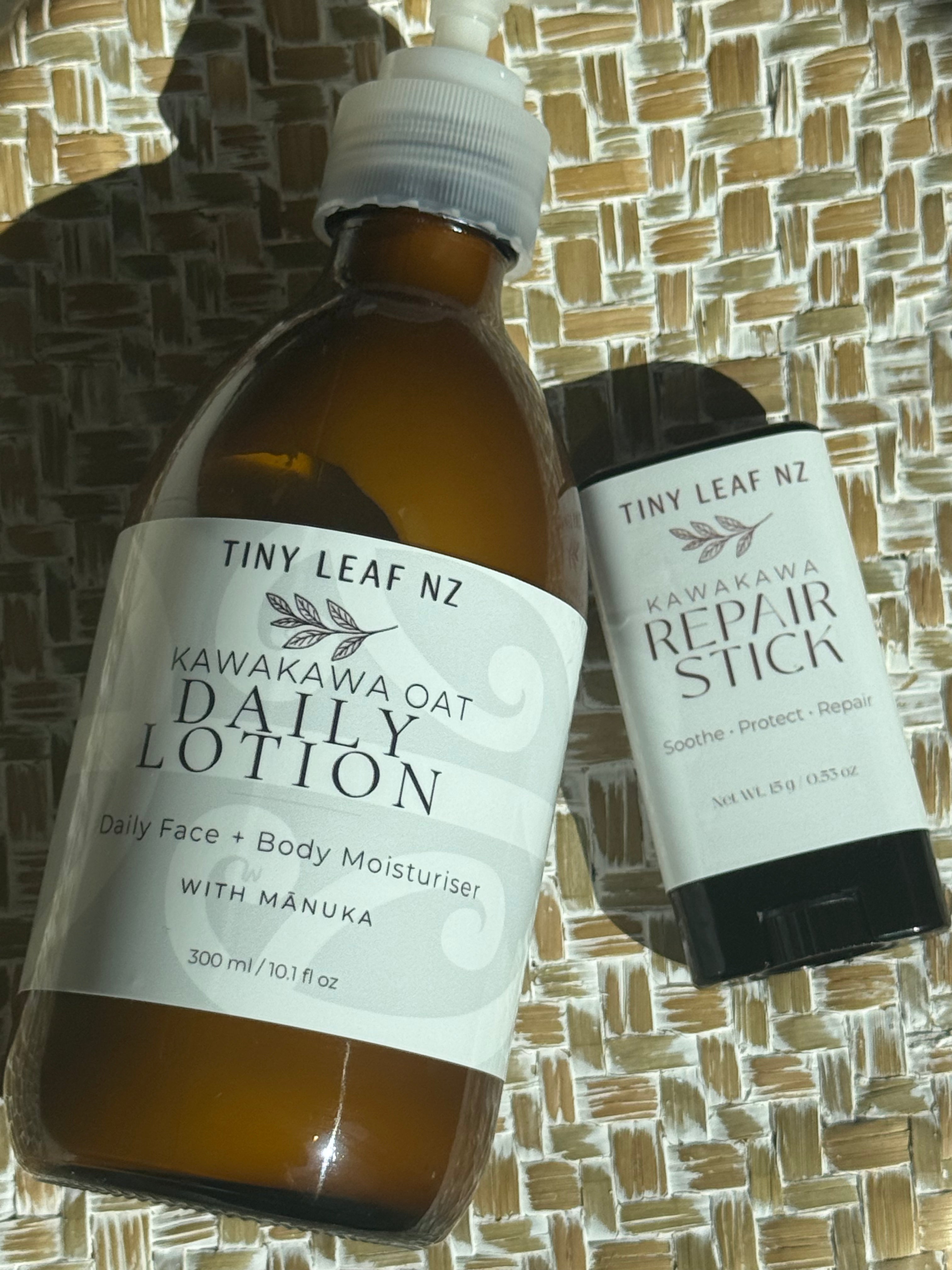 Tiny Leaf Nz Kawakawa Oat Daily Lotion in an amber glass bottle and Kawakawa Repair Stick in a black tube, both with white labels, displayed on a woven straw mat.