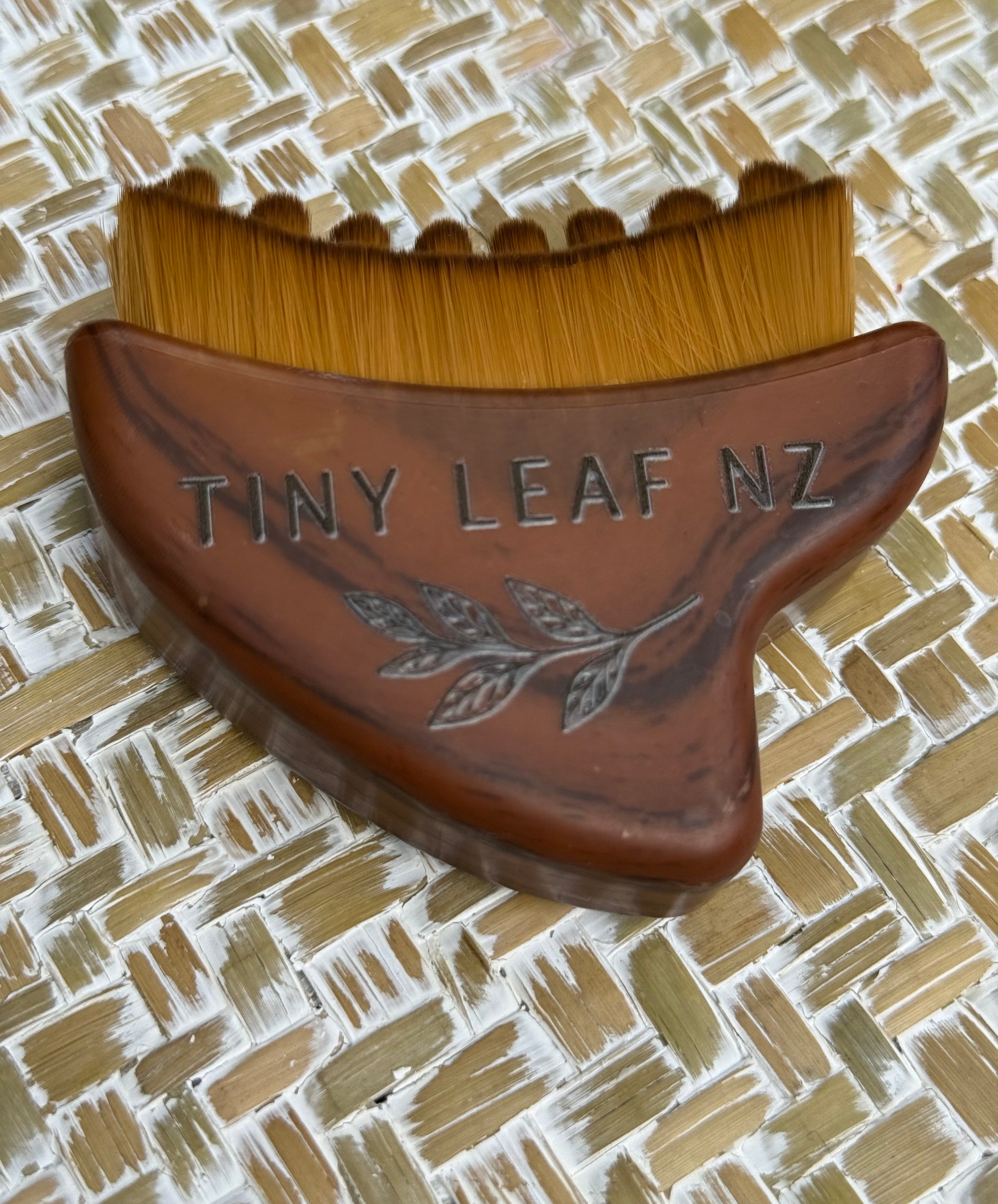 A wooden scalp massager with brown bristles and an engraved 'TINY LEAF NZ' logo, resting on a woven straw mat background.