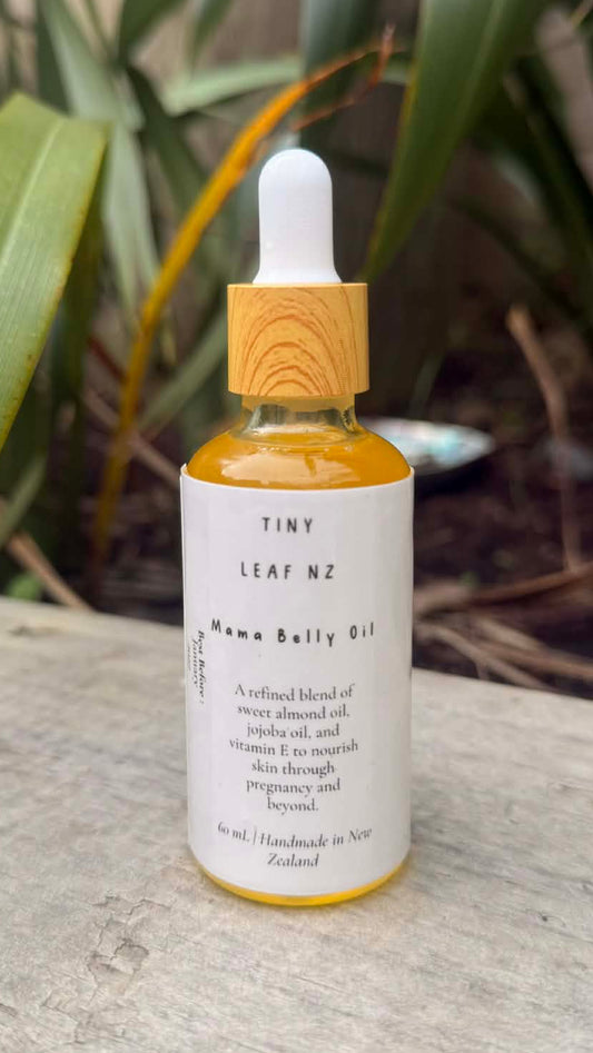 Bottle of TINY LEAF NZ Nuna Belly Oil with a dropper on a wooden surface with plants in the background.