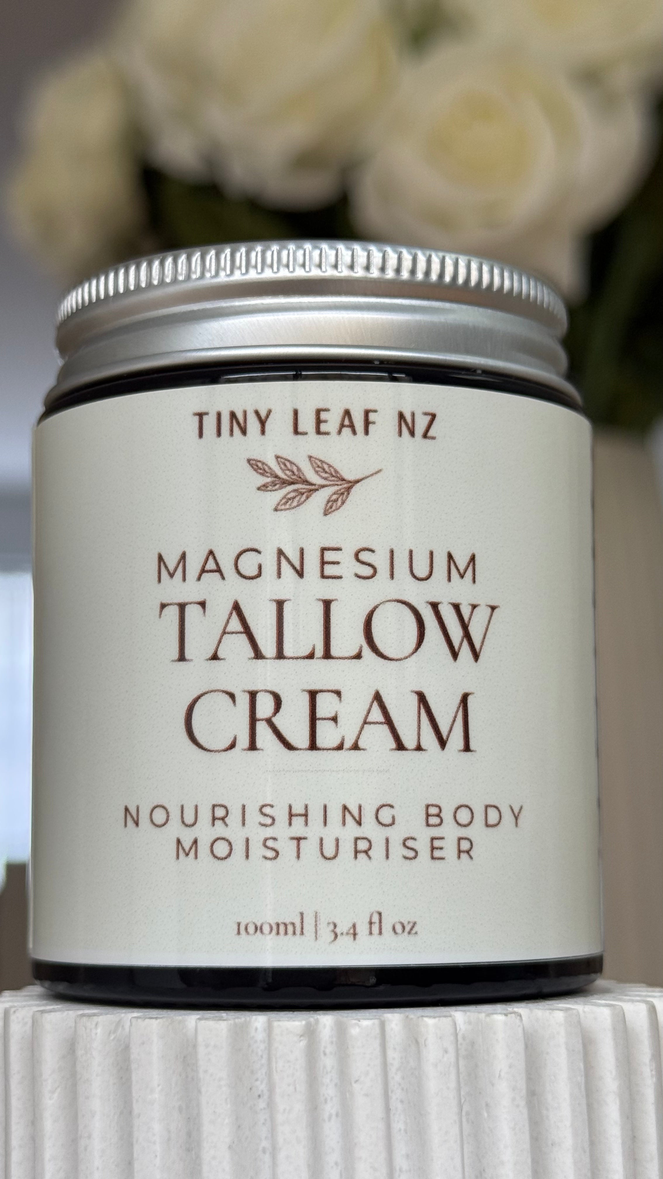 A jar of Tiny Leaf Nz Magnesium Tallow Cream body moisturizer, featuring a cream-colored label and a silver metal lid, set against a soft-focus background of white roses.
