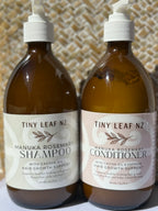 Two amber plastic pump bottles of Tiny Leaf Nz hair care products, labeled Manuka Rosemary Shampoo and Manuka Rosemary Conditioner, standing side by side against a woven beige background.