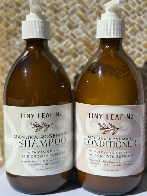 Two amber plastic pump bottles of Tiny Leaf Nz hair care products, labeled Manuka Rosemary Shampoo and Manuka Rosemary Conditioner, standing side by side against a woven beige background.