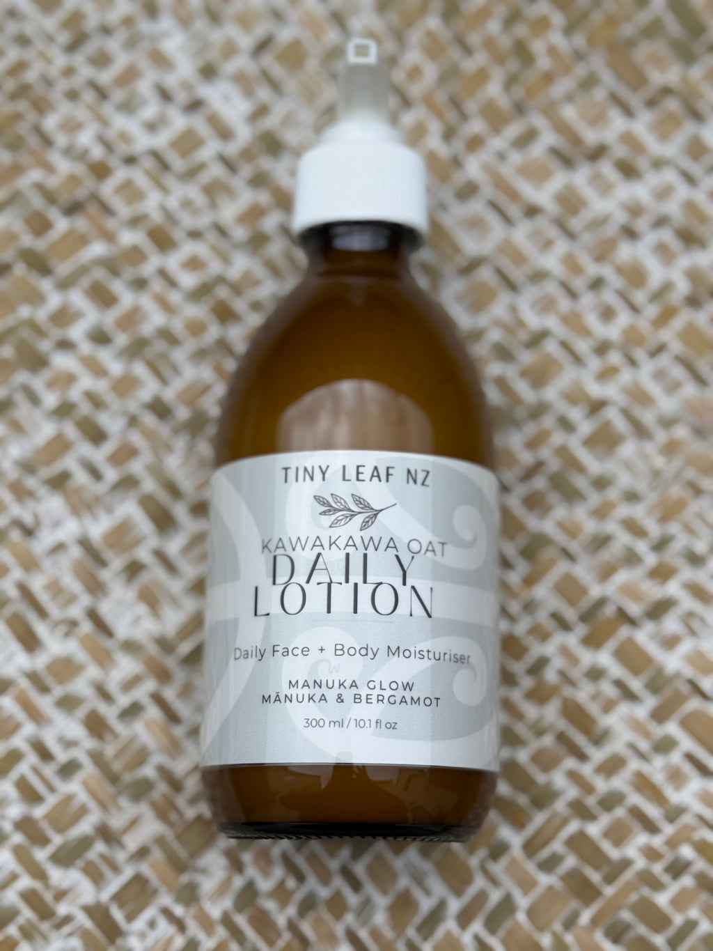 A 300ml amber glass bottle of Tiny Leaf Nz Kawakawa Oat Daily Lotion with a white cap and white label, placed on a textured beige woven mat background.