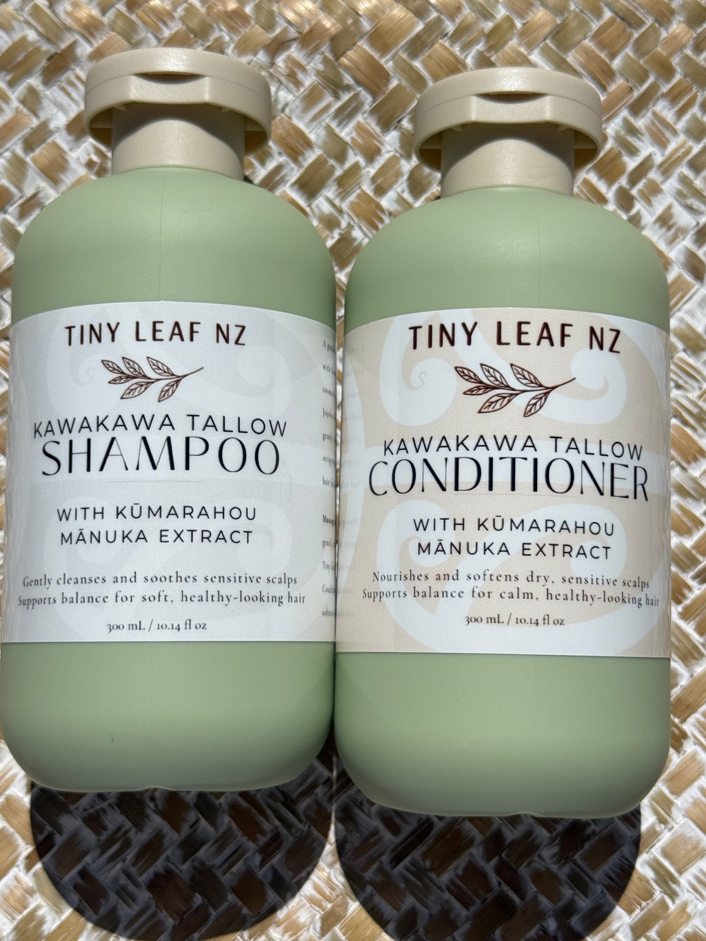 Two pale sage green bottles of Tiny Leaf NZ hair care products, labeled Kawakawa Tallow Shampoo and Kawakawa Tallow Conditioner, placed side by side on a woven mat background.